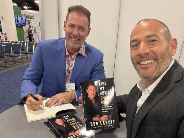 Rob Lancit seated at a table signing copies of Take My Advice, I'm Not Using It, a reader beside him