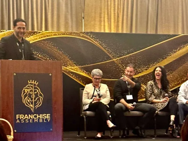 Rob Lancit seated on a Franchise Assembly panel, microphone in hand, pointing toward the audience, a gold and black backdrop behind