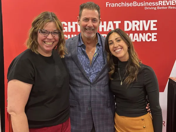 Rob Lancit between two women in front of a red Franchise Business Review wall