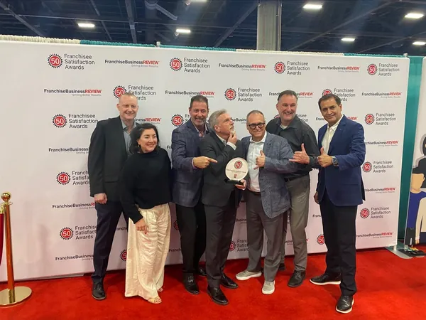 Six people on a red carpet in front of the Franchisee Satisfaction Awards backdrop, one holding an award
